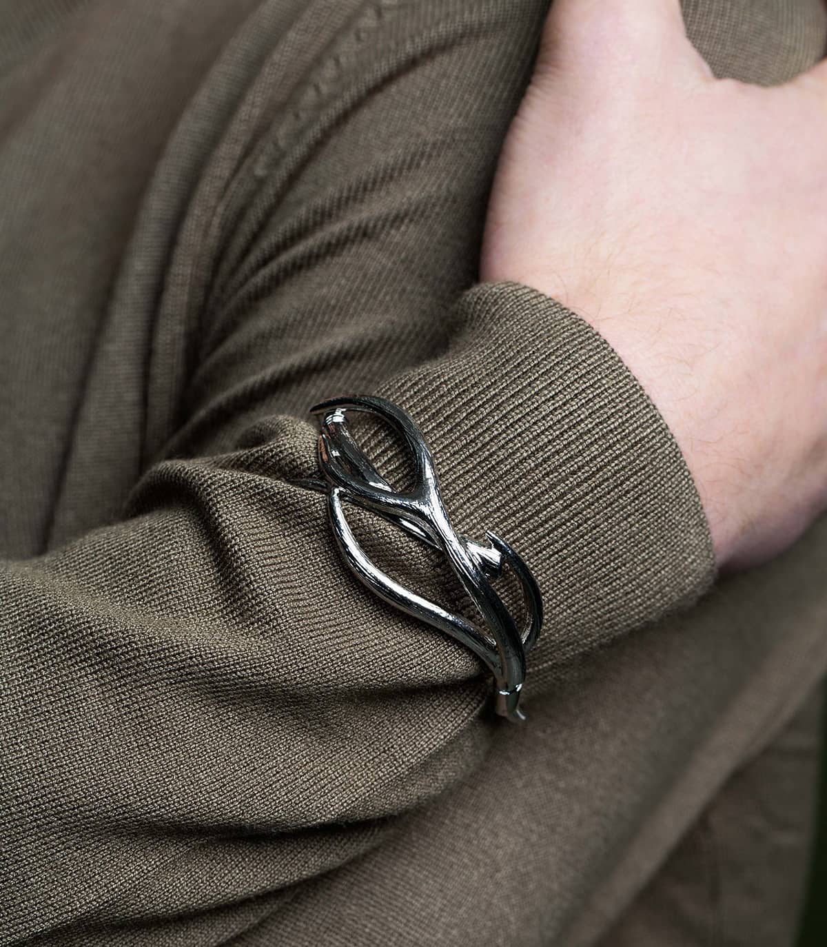 Zoom sur le poignet d'un homme portant un bracelet ruthénium inspiré de la forme des branches, sur les manches de son pull kaki.