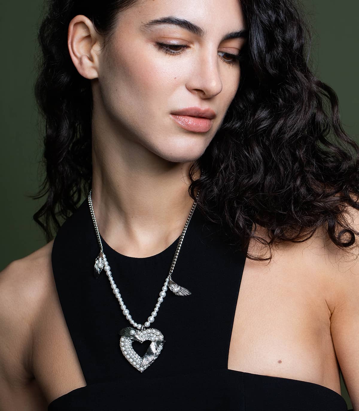 Femme portant au cou un long collier argenté agrémenté de perles, de strass, de petits motifs en forme de feuilles et d'un gros médaillon cœur.